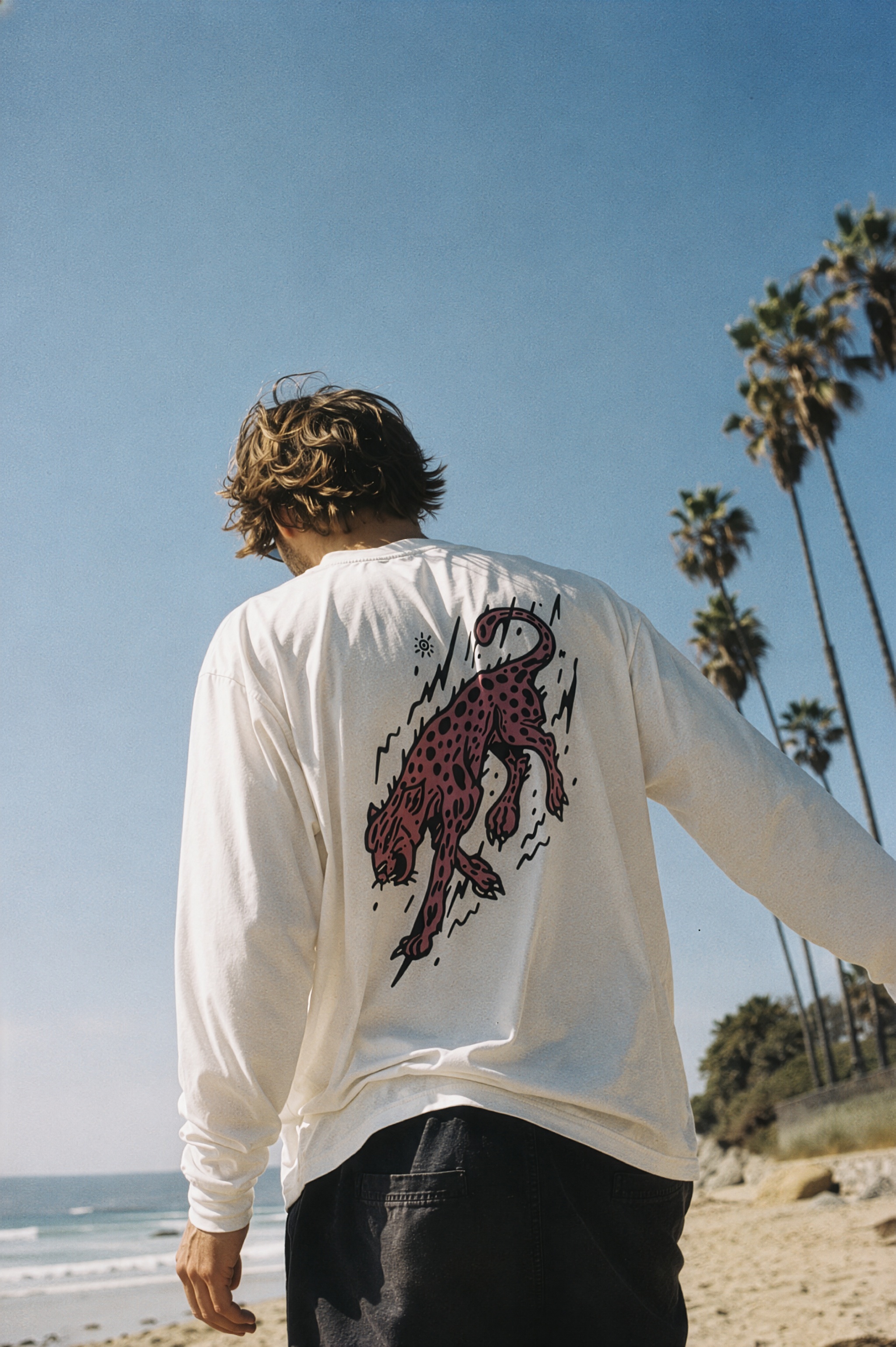 A man in a white long-sleeve rash guard, back to camera by the ocean, with the cheetah graphic in frame.