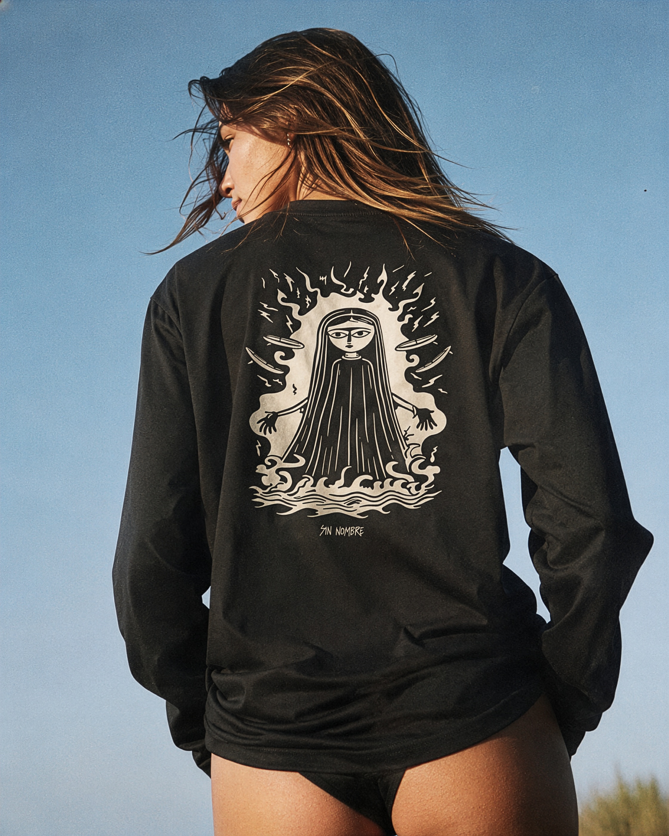 A woman in a black long-sleeve rash guard, back to camera against blue sky, with the sea-witch graphic in frame.